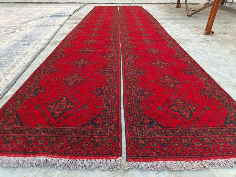 K&ouml;nnte beinhalten: Zwei lange, rechteckige rote Teppiche mit komplizierten geometrischen Mustern in Schwarz und Orange. Die Teppiche haben an den kurzen Enden einen Fransen. Die Teppiche liegen nebeneinander und scheinen handgefertigt zu sein.