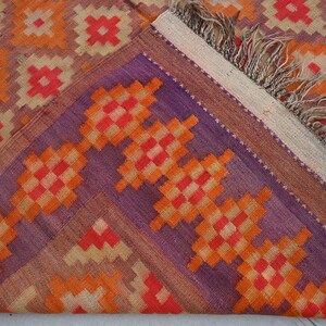 Op de afbeelding: Een close-up van een kleurrijk geweven tapijt met een geometrisch patroon in tinten van oranje, rood, paars en beige. Het tapijt heeft een franje rand.