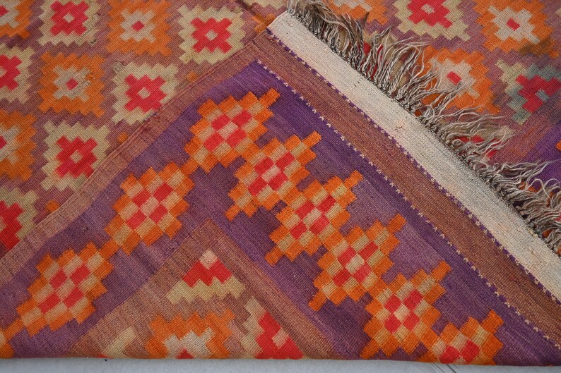 Op de afbeelding: Een close-up van een kleurrijk geweven tapijt met een geometrisch patroon in tinten van oranje, rood, paars en beige. Het tapijt heeft een franje rand.