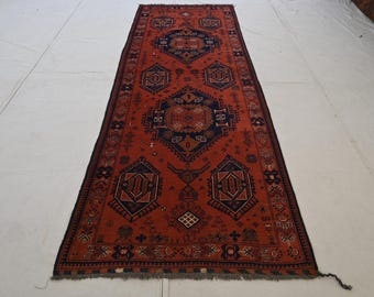 Puede incluir: Una alfombra larga y rectangular con un fondo rojo y intrincados patrones geométricos en azul marino y marrón. La alfombra presenta un diseño central con motivos repetidos y un borde oscuro. La alfombra es probablemente de lana y es adecuada para un pasillo o entrada.