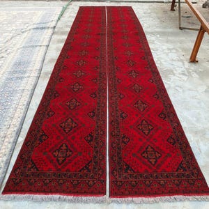 K&ouml;nnte beinhalten: Zwei lange, rechteckige rote Teppiche mit komplizierten geometrischen Mustern in Schwarz und Braun. Die Teppiche haben einen Fransen am unteren Rand und sind nebeneinander platziert. Die Teppiche liegen auf einer Betonoberfl&auml;che.