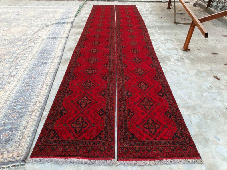 K&ouml;nnte beinhalten: Zwei lange, rechteckige rote Teppiche mit komplizierten geometrischen Mustern in Schwarz und Braun. Die Teppiche haben einen Fransen am unteren Rand und sind nebeneinander platziert. Die Teppiche liegen auf einer Betonoberfl&auml;che.