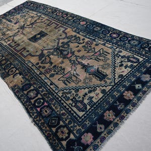 Peut inclure: Un tapis beige et bleu marine antique avec un motif g&eacute;om&eacute;trique complexe. Le tapis pr&eacute;sente un motif central avec des figures stylis&eacute;es et des motifs floraux, encadr&eacute; par une bordure bleu fonc&eacute; avec des formes et des accents floraux r&eacute;p&eacute;t&eacute;s. Les bords du tapis ont une finition frang&eacute;e.