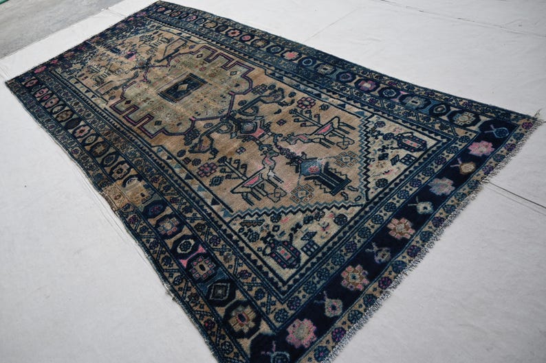 Peut inclure: Un tapis beige et bleu marine antique avec un motif g&eacute;om&eacute;trique complexe. Le tapis pr&eacute;sente un motif central avec des figures stylis&eacute;es et des motifs floraux, encadr&eacute; par une bordure bleu fonc&eacute; avec des formes et des accents floraux r&eacute;p&eacute;t&eacute;s. Les bords du tapis ont une finition frang&eacute;e.