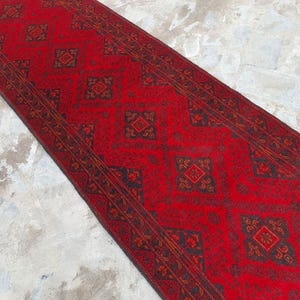 K&ouml;nnte beinhalten: Ein rechteckiger roter Teppich mit einem sich wiederholenden geometrischen Muster in Schwarz und Orange. Der Teppich liegt auf einer hellgrauen Oberfl&auml;che. Das Design des Teppichs zeigt Diamanten- und Quadratformen.