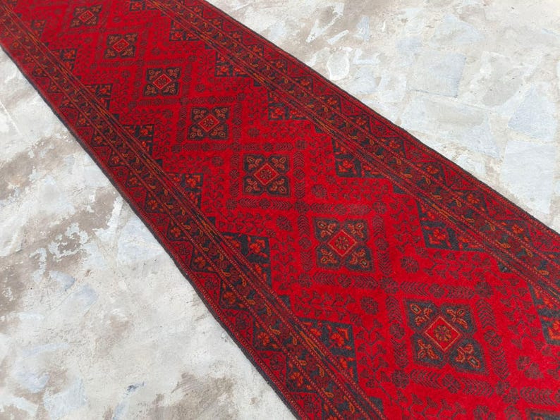 K&ouml;nnte beinhalten: Ein rechteckiger roter Teppich mit einem sich wiederholenden geometrischen Muster in Schwarz und Orange. Der Teppich liegt auf einer hellgrauen Oberfl&auml;che. Das Design des Teppichs zeigt Diamanten- und Quadratformen.