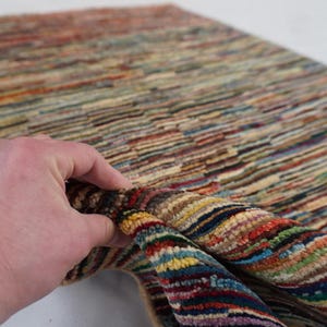 May include: A colorful rug with a striped pattern in shades of red, blue, yellow, and beige. The rug is being held up, revealing its folded edge. The rug's texture appears soft and inviting, suitable for home decor.