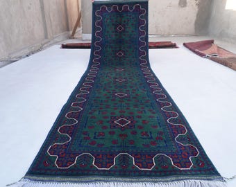 May include: A long, narrow runner rug with a green background and intricate floral and geometric patterns in shades of blue, red, and white. The rug has a fringe border.
