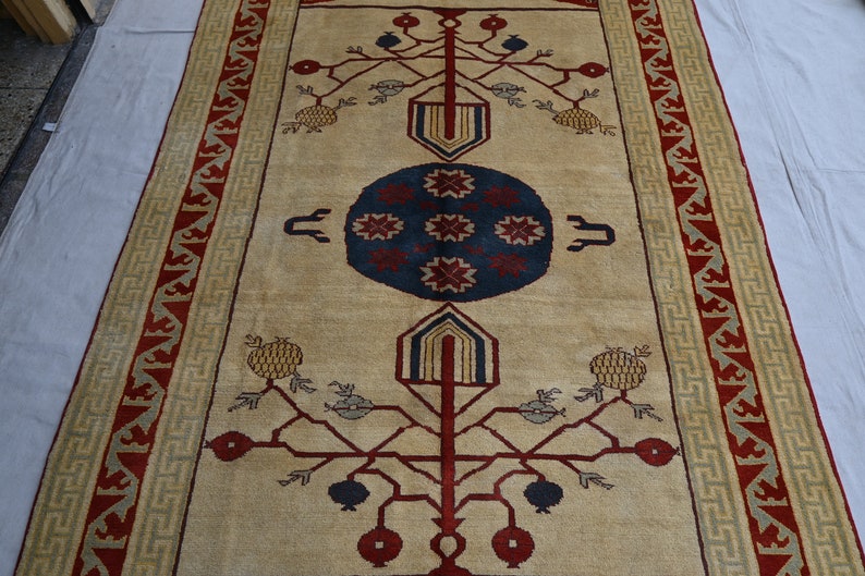 May include: A beige rug with a red, blue, and yellow geometric design. The rug features a central medallion with a stylized tree motif and pomegranate fruit.