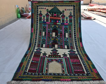 May include: A traditional prayer rug with a mosque design. The rug is beige with black, green, red, and blue accents. The mosque is surrounded by a border of geometric patterns.