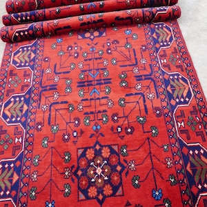 May include: A red runner rug with a geometric pattern in shades of blue, green, and brown. The rug has a fringe border and is rolled up.