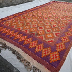 Op de afbeelding: Een kleurrijk tapijt met een geometrisch patroon in tinten oranje, rood en paars. Het tapijt heeft een franje rand en is geweven met een traditioneel ontwerp.