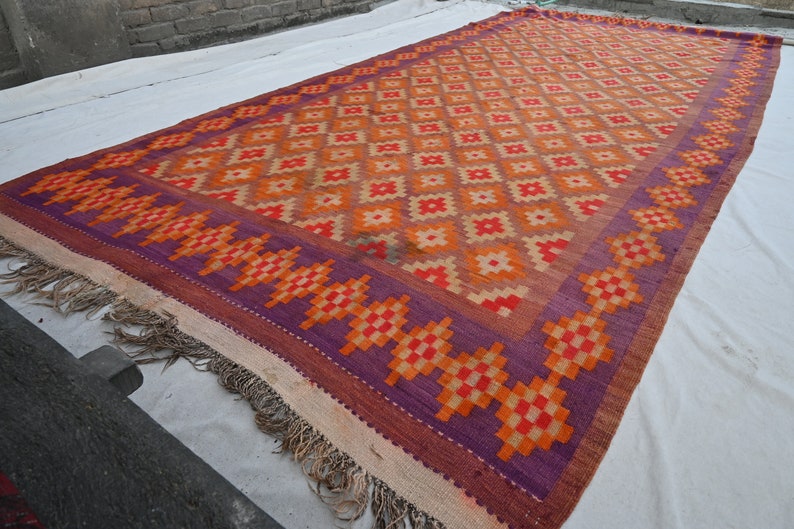 Op de afbeelding: Een kleurrijk tapijt met een geometrisch patroon in tinten oranje, rood en paars. Het tapijt heeft een franje rand en is geweven met een traditioneel ontwerp.