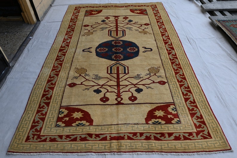 May include: A beige rug with a red and green border. The center of the rug features a stylized tree design with red and blue accents.