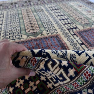 Pu&ograve; includere: Primo piano di un tappeto beige con intricati motivi geometrici in tonalit&agrave; di blu, verde e rosso. Il tappeto &egrave; piegato a met&agrave;, rivelando il lato inferiore con un motivo simile.