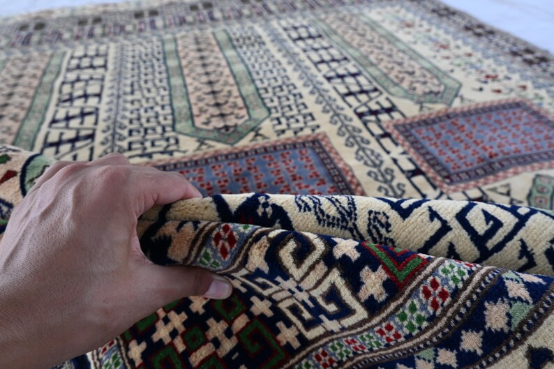 Pu&ograve; includere: Primo piano di un tappeto beige con intricati motivi geometrici in tonalit&agrave; di blu, verde e rosso. Il tappeto &egrave; piegato a met&agrave;, rivelando il lato inferiore con un motivo simile.