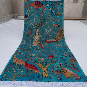 Op de afbeelding: Een rechthoekig, turquoise tapijt met een boom- en vogelontwerp. De boom is beige met groene bladeren, en de vogels zijn rood, oranje en geel. Het tapijt heeft een witte franje aan de onderkant.