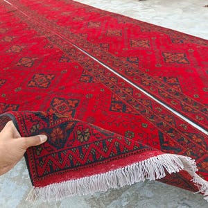 K&ouml;nnte beinhalten: Ein langer, roter Teppich mit komplizierten geometrischen Mustern in Marineblau und Braun. Der Teppich ist teilweise aufgerollt und zeigt einen detaillierten Rand und wei&szlig;e Fransen. Das Gesamtdesign ist traditionell und aufwendig.