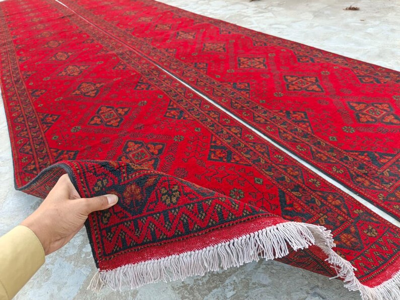 K&ouml;nnte beinhalten: Ein langer, roter Teppich mit komplizierten geometrischen Mustern in Marineblau und Braun. Der Teppich ist teilweise aufgerollt und zeigt einen detaillierten Rand und wei&szlig;e Fransen. Das Gesamtdesign ist traditionell und aufwendig.