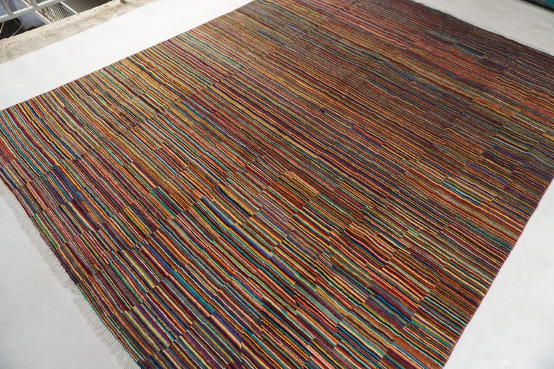 May include: A rectangular area rug featuring a dense pattern of thin, horizontal stripes in a vibrant palette of red, orange, yellow, blue, green, and dark brown. The rug has a flat-woven texture and includes short, light-colored fringe at the edges.