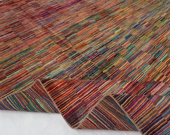May include: A rectangular area rug featuring a dense, multi-colored striped pattern composed of thin, horizontal lines in shades of red, orange, yellow, green, blue, and purple. The rug is folded at one corner, revealing the underside texture.