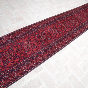 May include: A long, rectangular red runner rug with intricate geometric patterns and a dark blue border. The rug features a central design with repeating motifs and a fringed edge, suitable for a hallway or entryway.