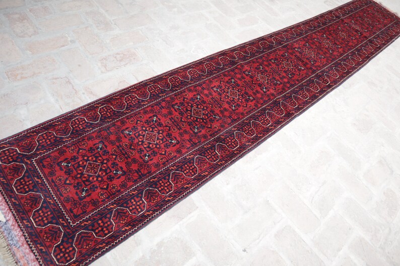 May include: A long, rectangular red runner rug with intricate geometric patterns and a dark blue border. The rug features a central design with repeating motifs and a fringed edge, suitable for a hallway or entryway.