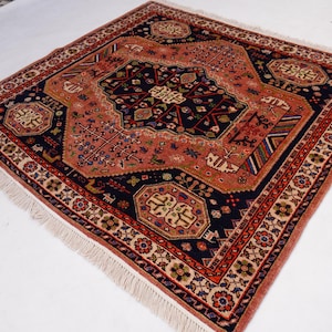 May include: Square, hand-knotted rug with a central medallion design. The rug features a navy blue field with a coral-colored central design and border. The border is detailed with floral and geometric patterns, and the rug has a fringe.