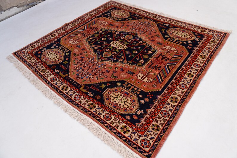 May include: Square, hand-knotted rug with a central medallion design. The rug features a navy blue field with a coral-colored central design and border. The border is detailed with floral and geometric patterns, and the rug has a fringe.