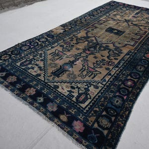 Peut inclure: Tapis rectangulaire avec un champ central beige orn&eacute; de motifs floraux et animaliers stylis&eacute;s en bleu, rose et noir. Le tapis a une bordure bleu fonc&eacute; avec des motifs g&eacute;om&eacute;triques r&eacute;p&eacute;titifs et des bords frang&eacute;s. Le tapis est pos&eacute; sur une surface blanche.