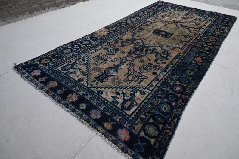 Peut inclure: Tapis rectangulaire avec un champ central beige orn&eacute; de motifs floraux et animaliers stylis&eacute;s en bleu, rose et noir. Le tapis a une bordure bleu fonc&eacute; avec des motifs g&eacute;om&eacute;triques r&eacute;p&eacute;titifs et des bords frang&eacute;s. Le tapis est pos&eacute; sur une surface blanche.