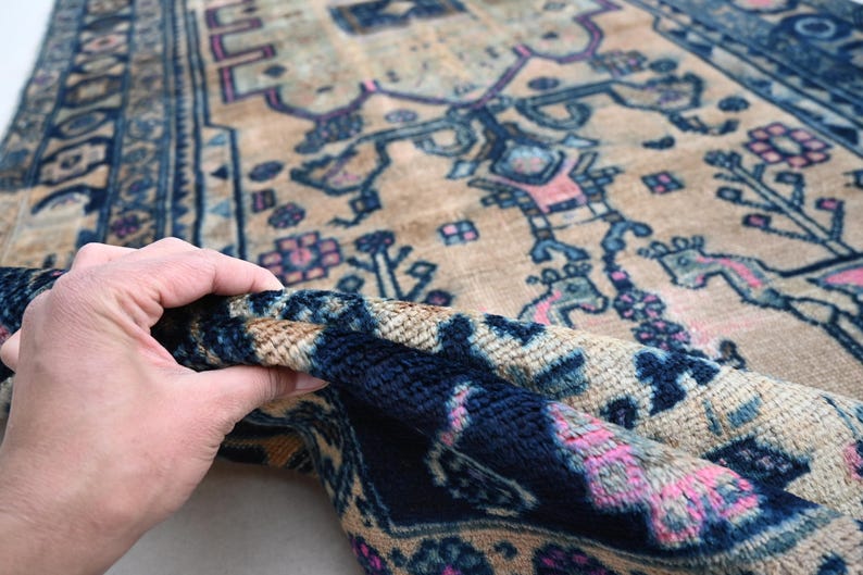 May include: A vintage rug with a beige background and intricate blue and pink floral patterns. The rug is folded in half, revealing a dark blue underside.