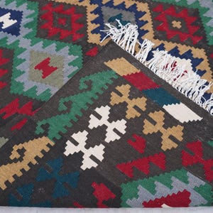 May include: A close-up of a colorful rug with a geometric pattern. The rug is made of wool and has a fringe border. The colors in the rug are red, green, blue, white, and brown.