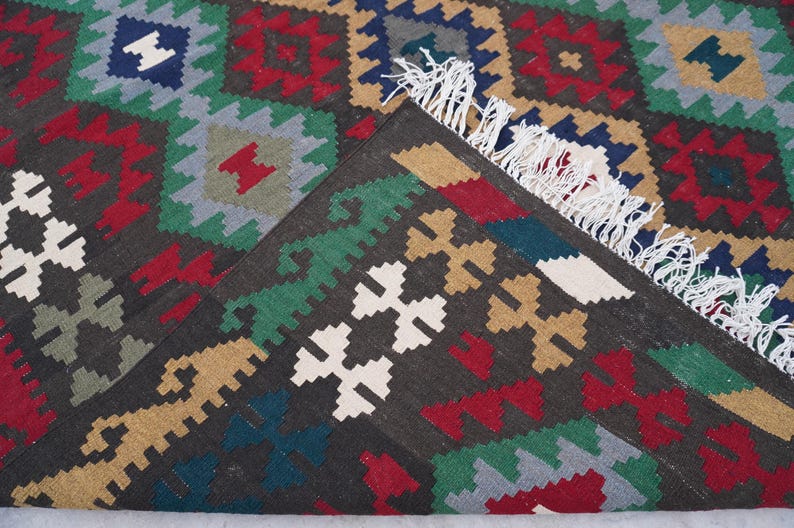 May include: A close-up of a colorful rug with a geometric pattern. The rug is made of wool and has a fringe border. The colors in the rug are red, green, blue, white, and brown.