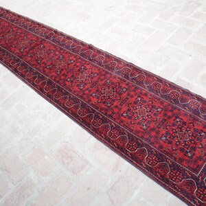 May include: A long, narrow red runner rug with intricate geometric patterns and a dark blue border. The rug's central field is a deep red, with repeating diamond and floral motifs. The rug is likely made of wool.