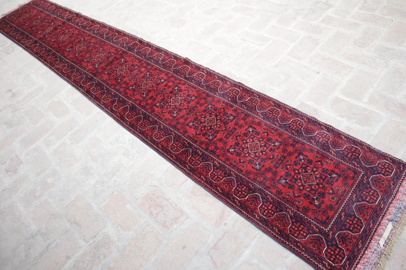 May include: A long, narrow red runner rug with intricate geometric patterns and a dark blue border. The rug's central field is a deep red, with repeating diamond and floral motifs. The rug is likely made of wool.