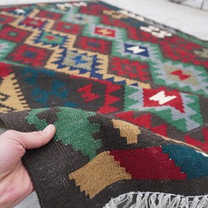 May include: A colorful hand-woven rug with a geometric pattern in shades of red, green, blue, and brown. The rug has a fringe border and is folded in half.