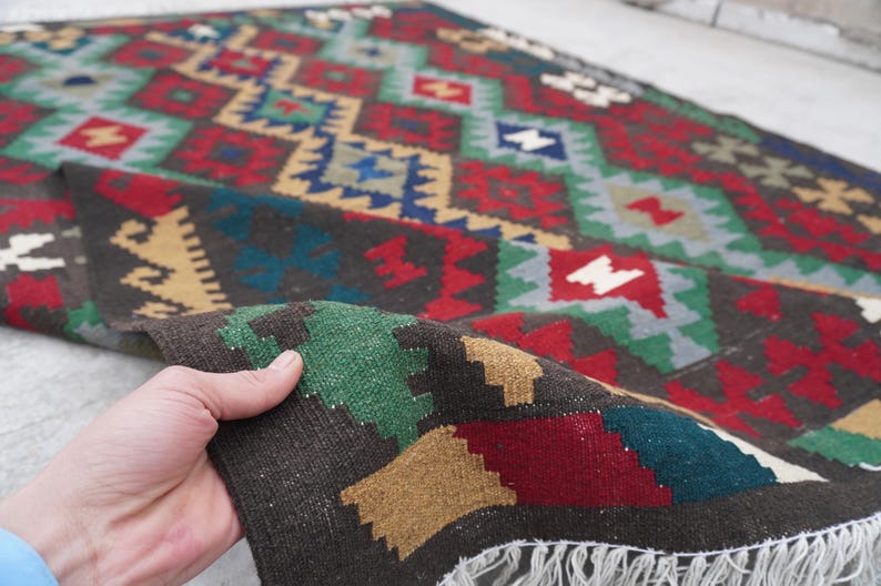 May include: A colorful hand-woven rug with a geometric pattern in shades of red, green, blue, and brown. The rug has a fringe border and is folded in half.