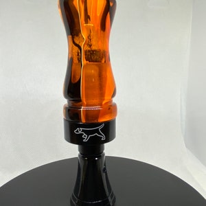 May include: A black and orange acrylic duck call with a black base and a white dog logo.