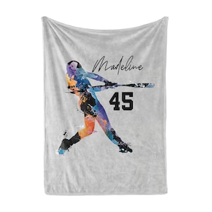 May include: Gray fleece blanket with a watercolor-style illustration of a baseball player in action. The player is wearing a blue and orange uniform with black accents. The text "Madeline 45" is printed on the blanket.