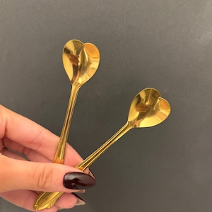 Könnte beinhalten: Zwei goldfarbene Löffel mit herzförmigen Laffen. Die Löffel haben lange, schlanke Stiele und werden vor einem dunklen Hintergrund gehalten. Die Löffel sind wahrscheinlich zum Servieren oder Essen gedacht.