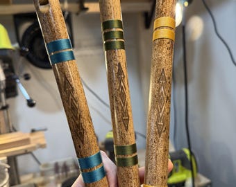 May include: Three wooden flutes with a speckled brown finish, each featuring a carved geometric arrow design and two sets of colored thread wraps. The left flute has teal wraps, the center has olive green wraps, and the right has yellow wraps.