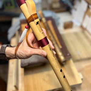May include: A handcrafted wooden flute with a natural finish, accented with burgundy bands and leather details. The flute features a decorative feather and bead, and is held in a person's hand. The instrument has several finger holes and engraved markings.