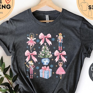 May include: Dark gray heather t-shirt with a Christmas-themed design featuring a nutcracker, a ballerina, a Christmas tree, and a gift box. The design is in watercolor style with pink bows and blue accents.