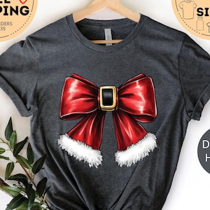 May include: A dark gray heather t-shirt with a red bow with a gold buckle and white fur trim. The shirt is hanging on a wooden hanger.
