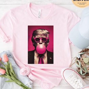 May include: Pink t-shirt with a graphic of a man with blonde hair wearing sunglasses and blowing a pink bubble. The man is wearing a dark suit and a pink tie.