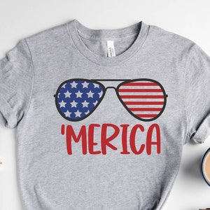 Puede incluir: Camiseta gris con un gr&aacute;fico de gafas de sol con un dise&ntilde;o de bandera estadounidense. El texto 'MERICA est&aacute; impreso debajo de las gafas de sol.