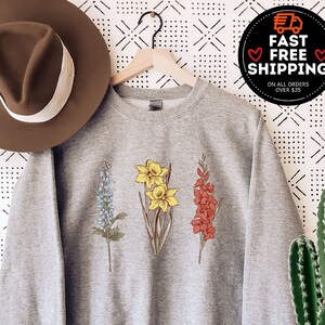 May include: A heather gray sweatshirt featuring embroidered botanical illustrations of blue, yellow, and red flowers. The sweatshirt is displayed on a hanger with a brown hat to the left. The image also includes a cactus in a white pot and text that says "MORE THAN 10 COLORS + SIZES".