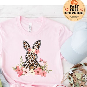 May include: Pink t-shirt with a leopard print bunny wearing a floral crown. The bunny is surrounded by pink flowers. The shirt is on a white wooden surface with a pair of blue denim shorts, a blue baseball cap, white sneakers, and a floral wreath.