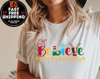 May include: White t-shirt with the word "Believe" in colorful, festive lettering. The design incorporates Christmas-themed elements like a Santa hat, snowflake, and reindeer antlers. The shirt is paired with blue jeans.